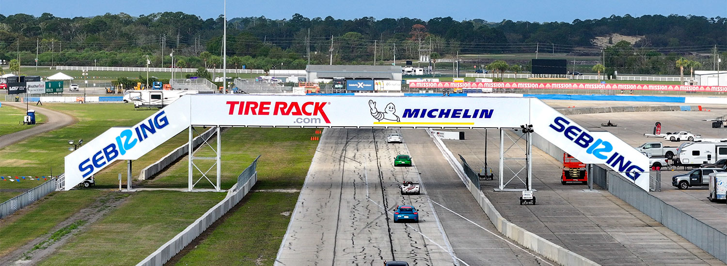 Multiple race cars travel down the front straight at Sebring International Raceway, passing beneath a pedestrian bridge displaying Tire Rack and Michelin branding, with “Sebring” written along both sides.
