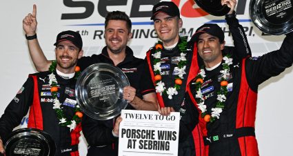 Four men wearing matching black and red Porsche racing team uniforms stand together on a podium, smiling and celebrating a victory. Two of them hold large circular trophies, while another raises one overhead. The group wears black caps and floral victory garlands around their necks. One person in the center holds a sign that reads “Porsche Wins at Sebring” with “Twelve to Go” above it. The backdrop behind them features racing event branding, including references to Sebring. The mood is celebratory, with the team posing closely together and looking toward the camera.