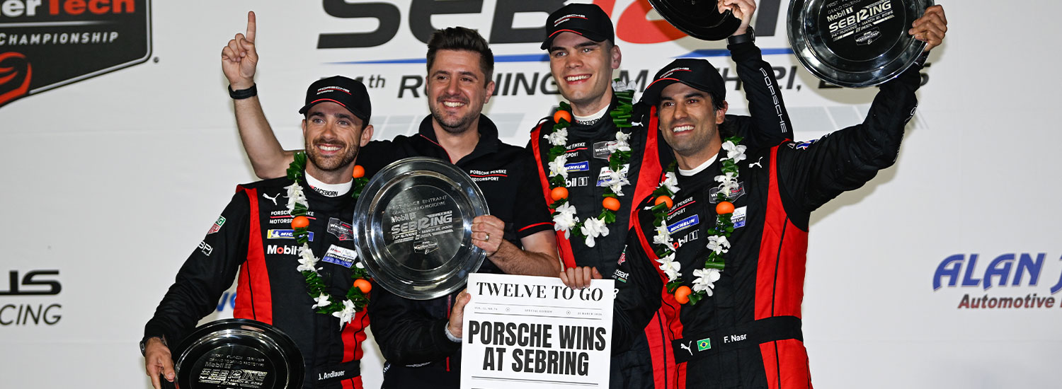 Four men wearing matching black and red Porsche racing team uniforms stand together on a podium, smiling and celebrating a victory. Two of them hold large circular trophies, while another raises one overhead. The group wears black caps and floral victory garlands around their necks. One person in the center holds a sign that reads “Porsche Wins at Sebring” with “Twelve to Go” above it. The backdrop behind them features racing event branding, including references to Sebring. The mood is celebratory, with the team posing closely together and looking toward the camera.
