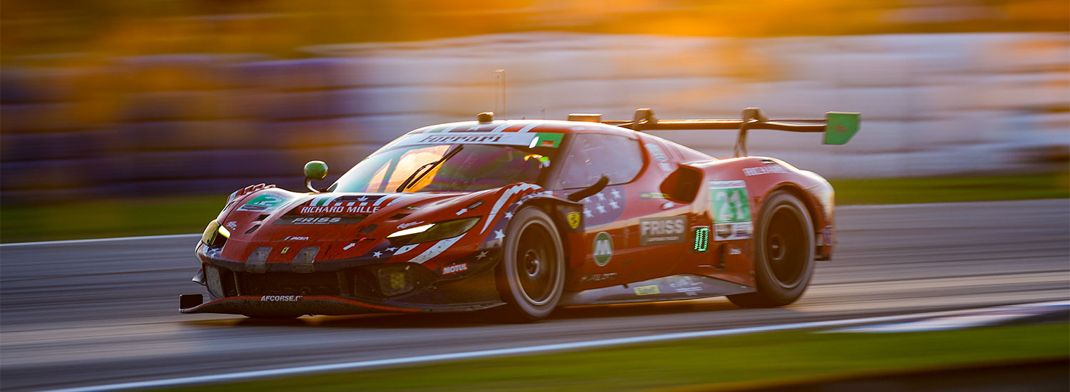 A red GT-style endurance race car, marked with the number 21 and covered in sponsor logos, speeds along a racetrack. The car is captured in motion with strong horizontal blur in the background, emphasizing its high speed. The lighting is warm and golden, suggesting sunset or early evening, with sunlight reflecting off the car’s body and windshield. The track surface appears smooth, and the surrounding barriers and environment are blurred due to the panning effect of the camera. The car has a low, aerodynamic profile with a large rear wing and wide tires designed for racing performance.