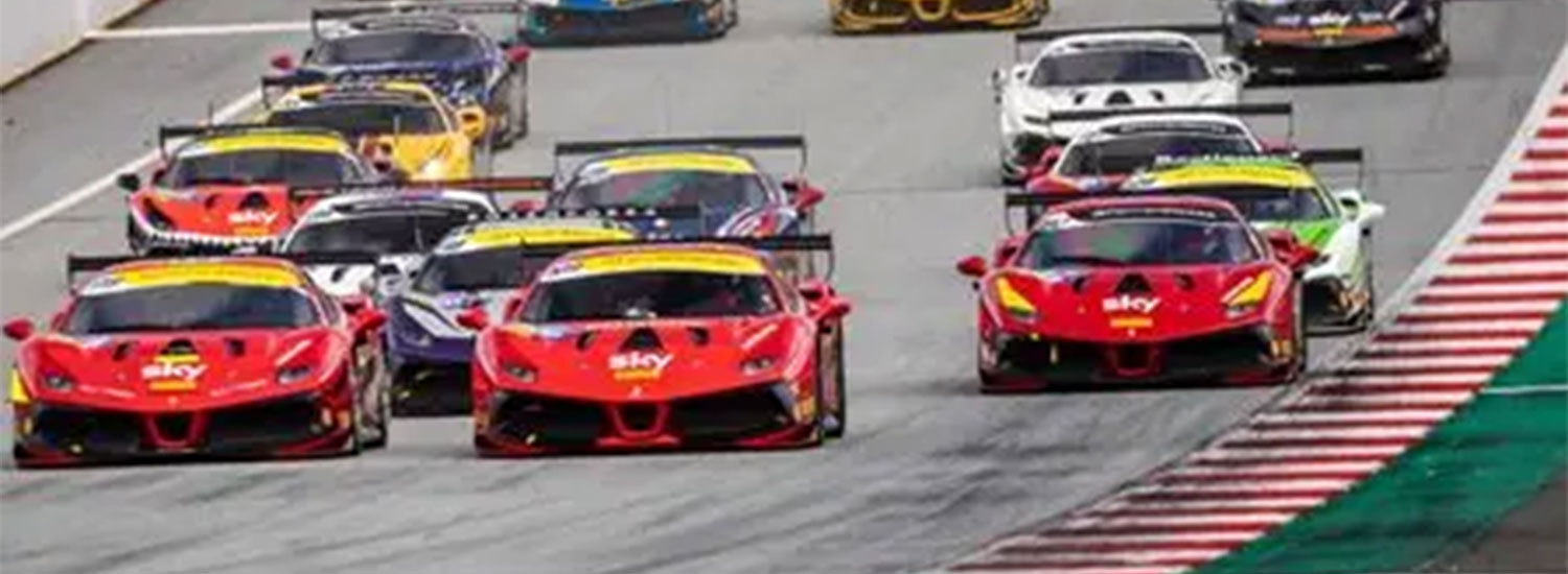 Multiple Ferrari race cars, primarily red with sponsor markings, race side-by-side in a tight group through a corner at a road course, with red-and-white curbing and track runoff visible during a Ferrari Challenge race.
