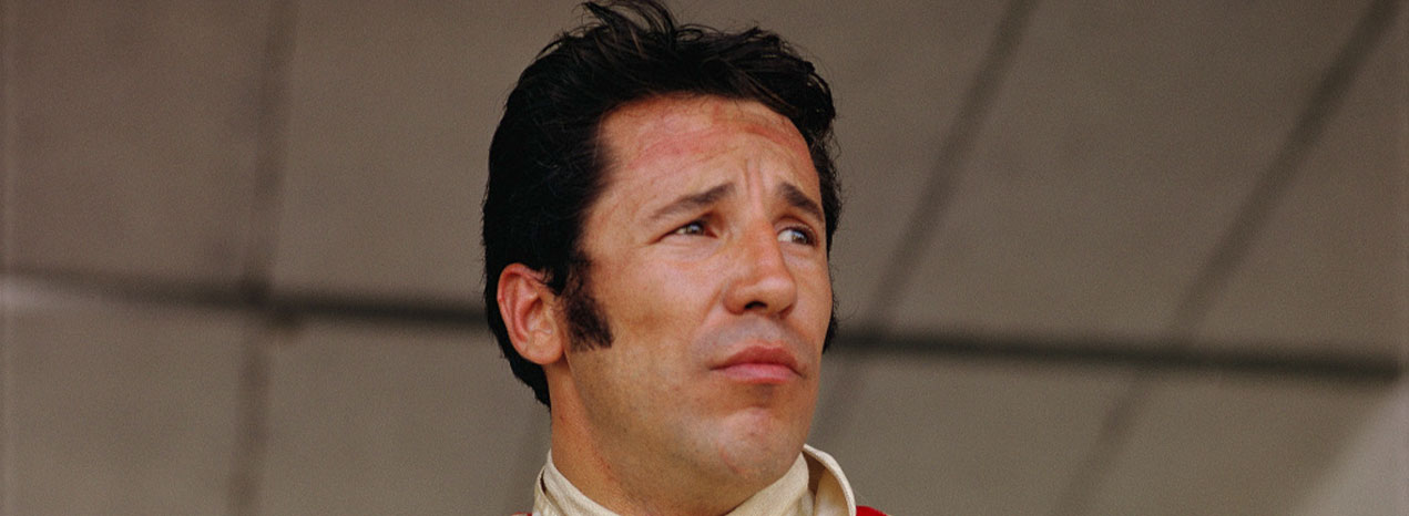 Close-up portrait of Mario Andretti in a racing suit, looking upward against a neutral background.